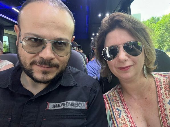 Two adults wearing sunglasses pose for a selfie on a tour bus, seated among other passengers with bus seats and green trees visible through the window.