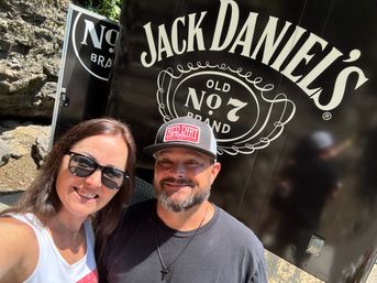 Outdoor selfie of a smiling couple in front of a black trailer featuring a large white vintage liquor-style logo, with rocky cliff background and bright sunlight.