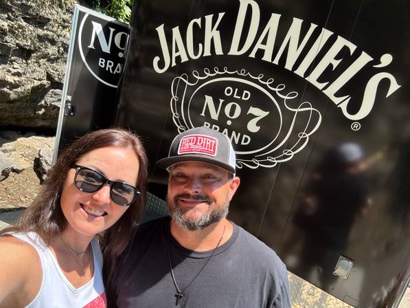 Outdoor selfie of a smiling couple in front of a black trailer featuring a large white vintage liquor-style logo, with rocky cliff background and bright sunlight.