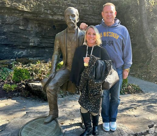 Two adults smiling and posing beside a seated bronze statue with one foot on a barrel on a rocky outdoor path; woman in a black poncho holding a cup and leopard-print coat, man in a blue hoodie and jeans standing behind her on a sunny day.