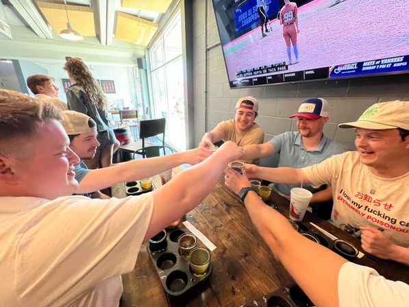 Group of friends toasting with small beer glasses over a wooden table and beer flight trays inside a bright craft brewery/sports bar with large windows and a TV showing a softball game.