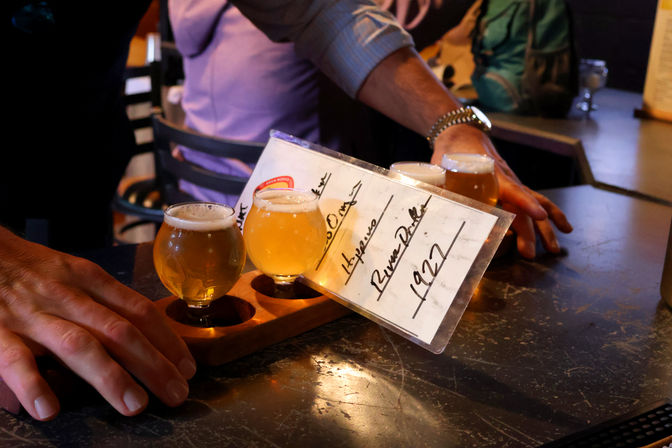 Flight of craft beers on a wooden paddle with small tulip tasting glasses and a handwritten label, hands on a worn bar counter in a cozy pub setting