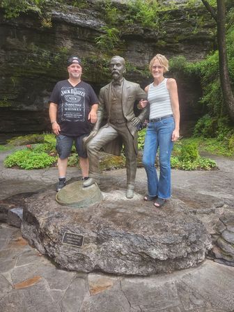 Two visitors posing beside a bronze bearded gentleman statue on a rock platform in a lush, mossy canyon park with layered stone cliffs and green plants