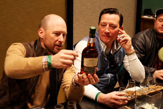Two men in an indoor tasting room inspecting a whiskey bottle during a whiskey tasting, with tasting glasses and wooden flight boards on the table.