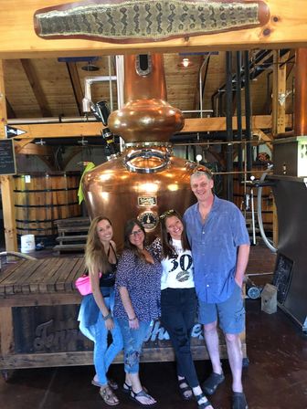 Four smiling adults posing in front of a large copper pot still inside a rustic craft distillery tasting room with wooden beams and barrels.