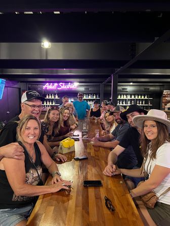 Large group of friends smiling and gathered along a long wooden bar in a dimly lit lounge with a neon sign advertising slushie cocktails and rows of bottles behind them, wearing casual hats and enjoying a festive night out.