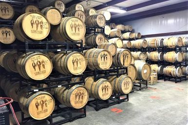Rows of oak barrels stacked on black metal racks inside a bright distillery warehouse, wooden barrel heads stamped with designs, concrete floor and industrial lighting