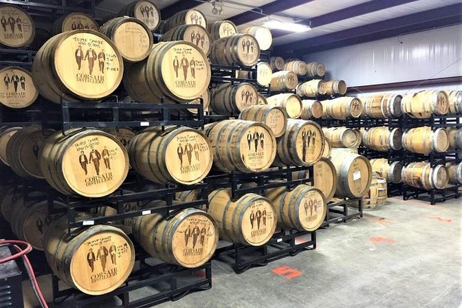 Rows of oak barrels stacked on black metal racks inside a bright distillery warehouse, wooden barrel heads stamped with designs, concrete floor and industrial lighting