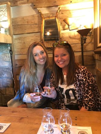 Two smiling women toasting whiskey in Glencairn glasses at a rustic wood‑paneled tasting room, seated at a wooden bar beneath a vintage mirror and warm brass lamp — cozy distillery vibes.