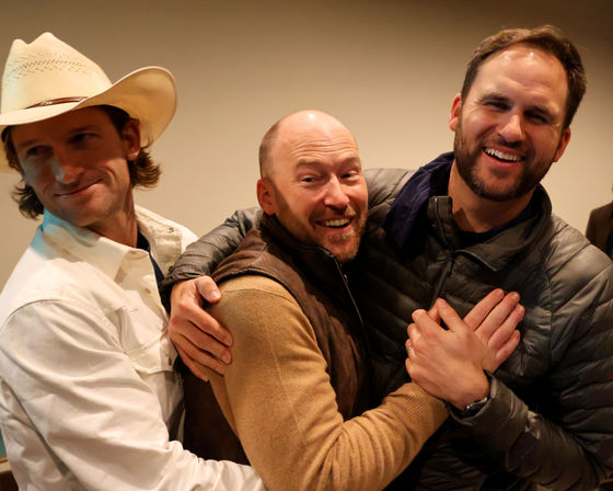 Three laughing men at a casual indoor gathering sharing a friendly group hug — left man in a cowboy hat and white shirt, center man in a brown vest, right man in a gray puffer jacket.