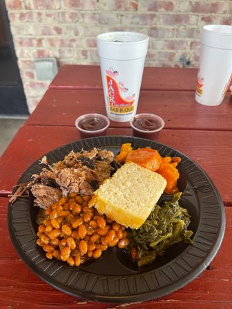 Classic Southern barbecue plate with pulled pork, baked beans, collard greens, cornbread and candied sweet potatoes on a black disposable plate, next to a white takeout cup on a red picnic table by a brick wall