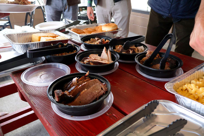 Mouthwatering BBQ spread on a red picnic table — plastic bowls of sliced smoked brisket and pulled pork, foil trays of burger buns and mac and cheese, and tongs for a self-serve outdoor buffet.