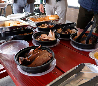 Mouthwatering BBQ spread on a red picnic table — plastic bowls of sliced smoked brisket and pulled pork, foil trays of burger buns and mac and cheese, and tongs for a self-serve outdoor buffet.