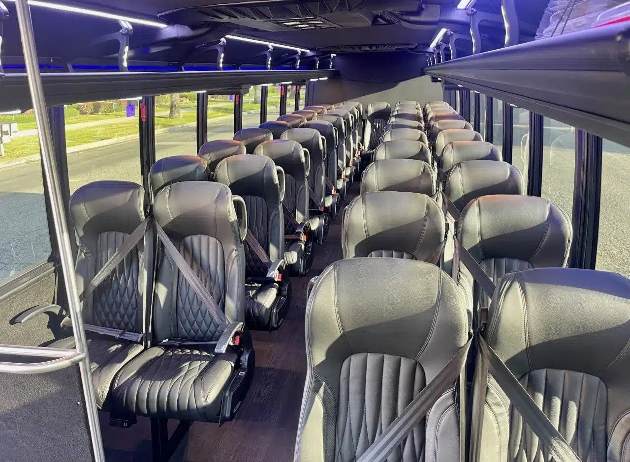 Empty luxury shuttle bus interior with rows of black leather seats with seatbelts, center aisle, overhead luggage racks, LED accent lighting and large windows