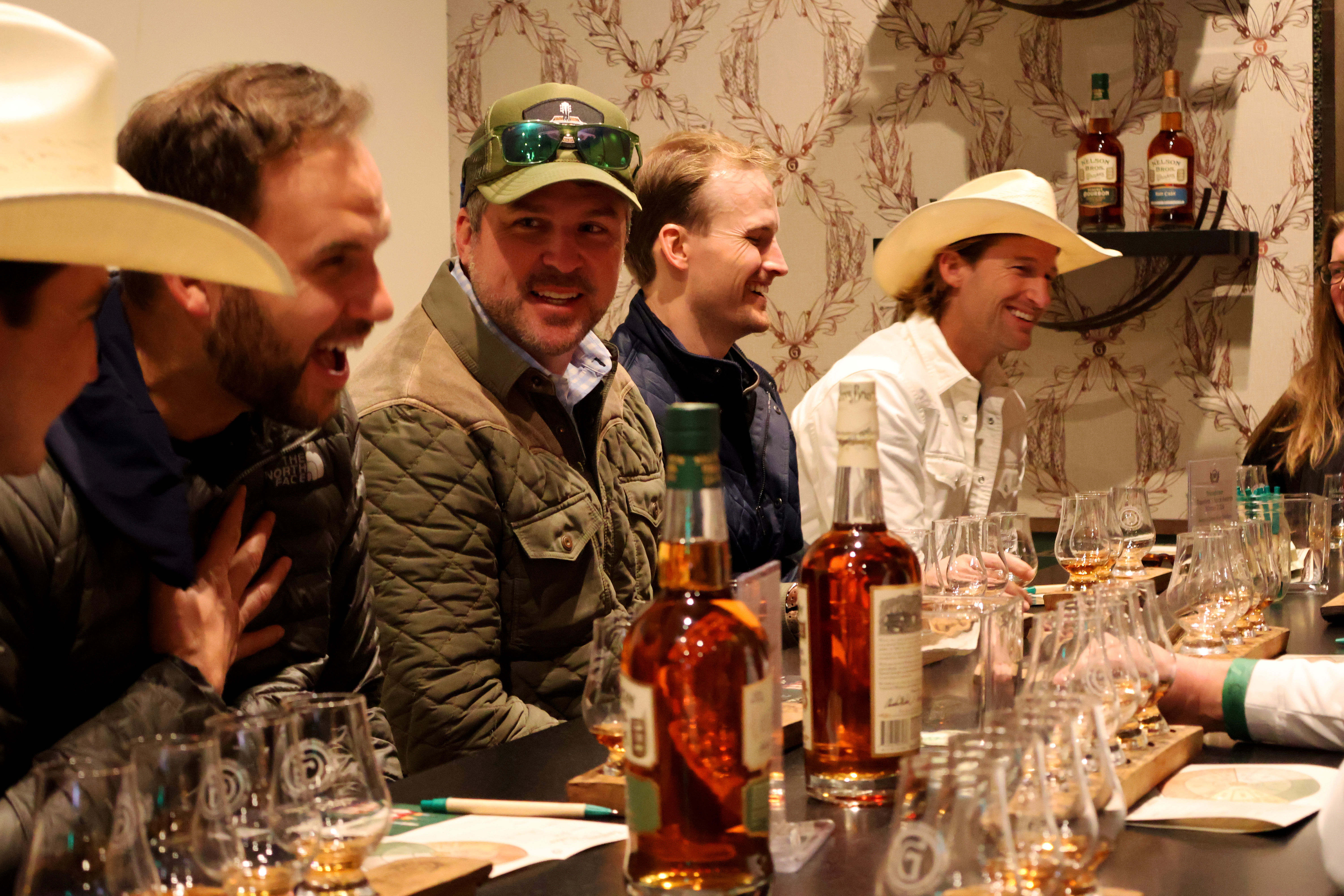 Smiling group in cowboy hats and jackets gathered at a bar for a whiskey tasting flight, with amber bottles and rows of tasting glasses on the counter in a warm indoor setting.