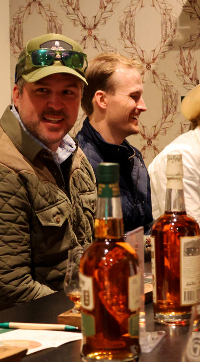 Smiling group in cowboy hats and jackets gathered at a bar for a whiskey tasting flight, with amber bottles and rows of tasting glasses on the counter in a warm indoor setting.
