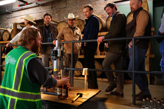 Worker in a green safety vest bottling whiskey while visitors in jackets and a cowboy hat watch inside a craft distillery with oak barrels stacked against brick walls.