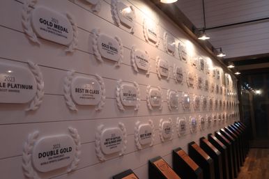 Tasting-room interior with a long wall of 2023 award plaques and laurel-style medals under hanging lights, wooden floor and angled display stands.