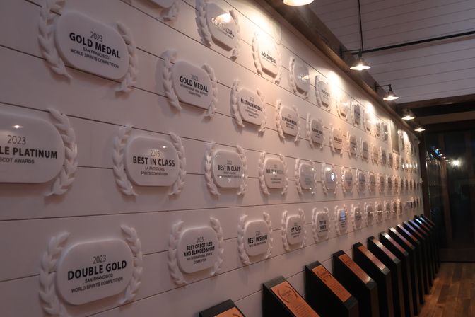 Tasting-room interior with a long wall of 2023 award plaques and laurel-style medals under hanging lights, wooden floor and angled display stands.