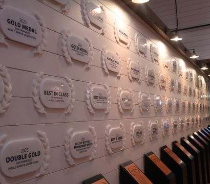 Tasting-room interior with a long wall of 2023 award plaques and laurel-style medals under hanging lights, wooden floor and angled display stands.