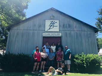 Group of adults posing on steps in front of a weathered barn-style distillery building with a painted logo above the door, bright blue sky and green shrubs.