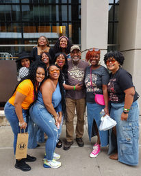 Cheerful group of adults posing for a group photo on a downtown sidewalk in front of a modern glass building, wearing casual denim, bright sneakers and sunglasses, laughing and hugging.