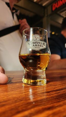 Close-up of a whiskey tasting glass filled with a golden amber spirit on a polished wooden bar, with blurred patrons and warm taproom lighting in the background.