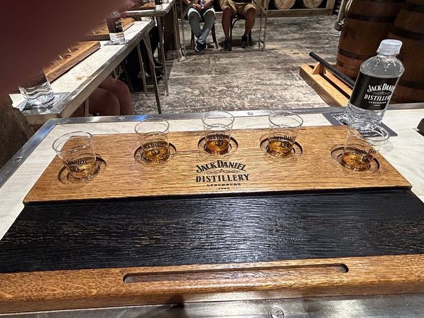Wooden whiskey tasting flight holding five small sample glasses on a metal table in a rustic distillery tasting room, with oak barrels and a water bottle visible in the background.