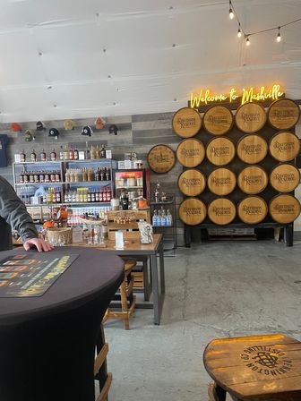 Interior of a Nashville tasting room with stacked wooden barrels, neon 'Welcome to Nashville' sign, shelves of bottled spirits, merch hats, tasting tables and stools