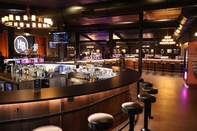 Industrial-chic cocktail lounge interior at night with a large U-shaped backlit bar, rows of round black stools, bottles and glassware on the counter, chandeliers and a small stage in the background.