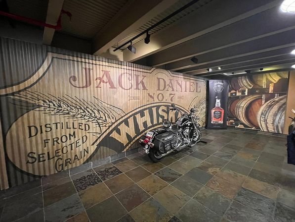 Black motorcycle parked in a dim whiskey tasting room with a large vintage whiskey mural on a corrugated wall and stacked oak barrels