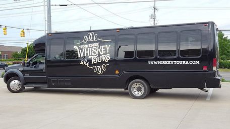 Black passenger shuttle bus parked in a suburban lot, featuring vintage-style whiskey tours lettering and a Tennessee motif on its side.