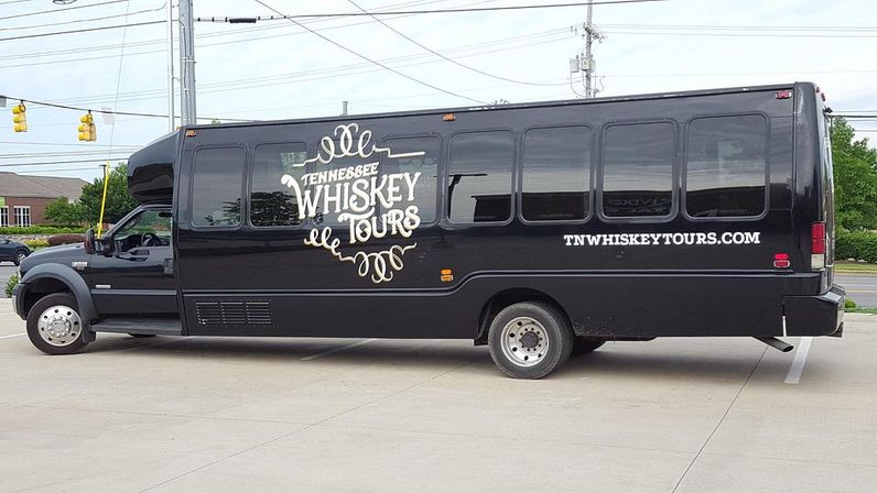 Black passenger shuttle bus parked in a suburban lot, featuring vintage-style whiskey tours lettering and a Tennessee motif on its side.