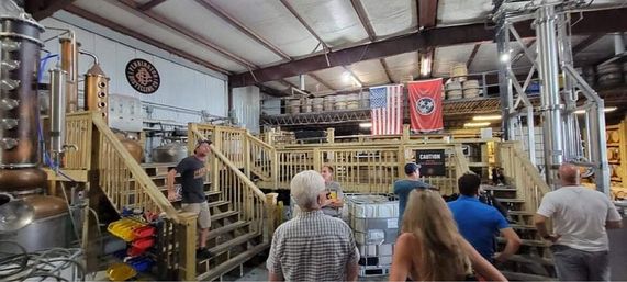 Distillery tour inside an industrial warehouse with copper stills, stacked barrels and a wooden mezzanine; visitors stand near equipment beneath an American and state flag.