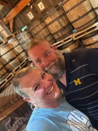 Cheerful couple selfie inside a rustic barrel room, smiling in front of stacked wooden aging barrels on pallets — man with a gray beard in a navy polo with a yellow 'M' logo and woman in a light blue t-shirt.