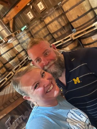 Cheerful couple selfie inside a rustic barrel room, smiling in front of stacked wooden aging barrels on pallets — man with a gray beard in a navy polo with a yellow 'M' logo and woman in a light blue t-shirt.
