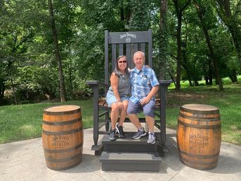 Smiling couple sitting on an oversized wooden branded rocking chair flanked by two wooden whiskey barrels on a sunny, tree-lined lawn.