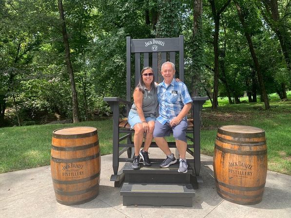 Smiling couple sitting on an oversized wooden branded rocking chair flanked by two wooden whiskey barrels on a sunny, tree-lined lawn.