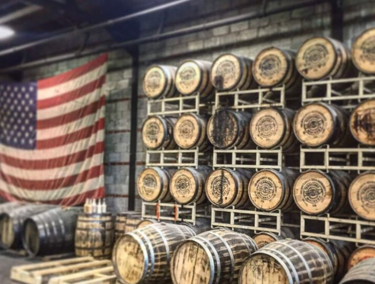 Rustic distillery warehouse interior with rows of stacked oak whiskey barrels on metal racks, loose barrels on pallets, and a large American flag hanging on a brick wall.