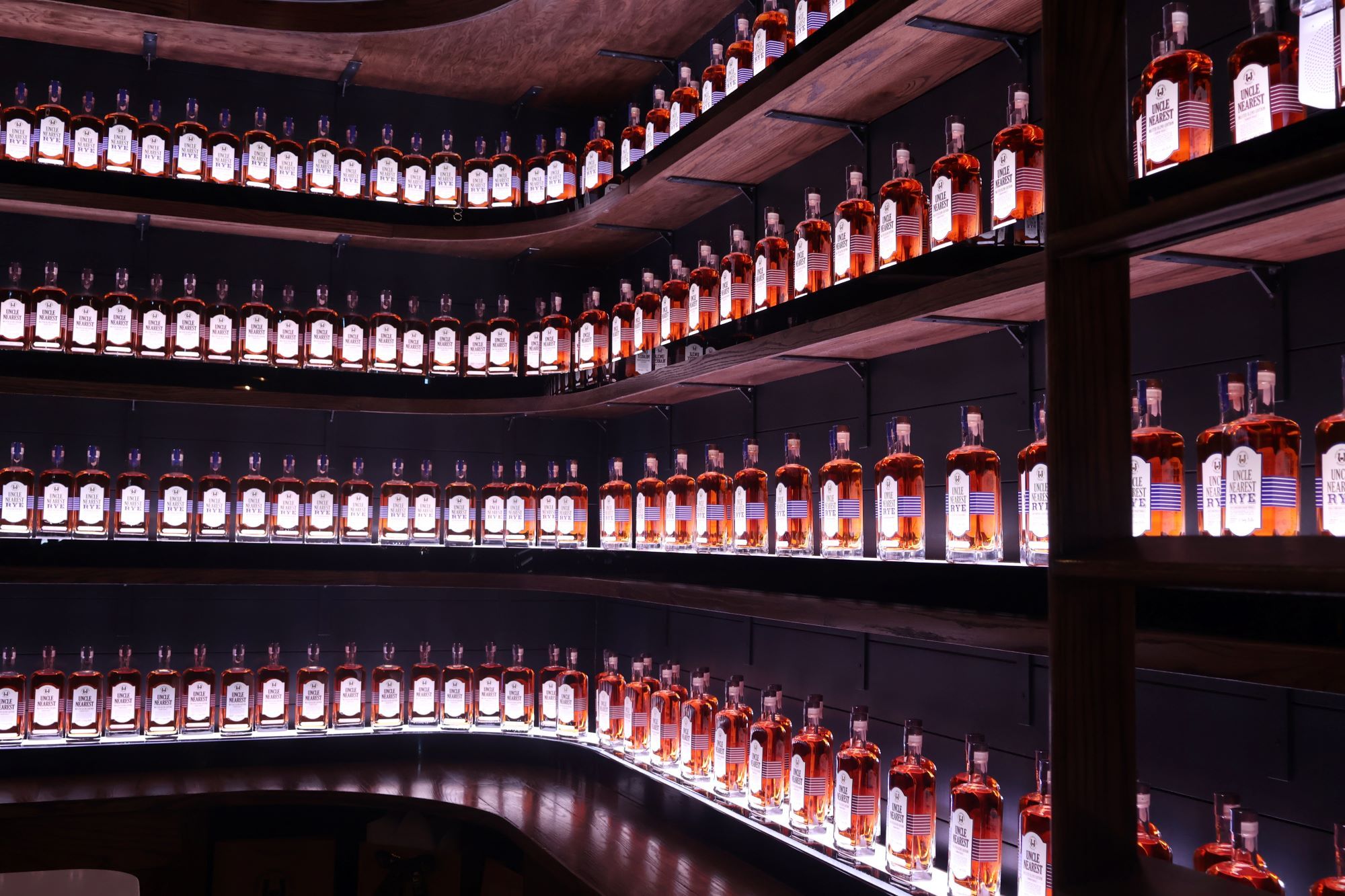 Curved wooden shelves lined with dozens of backlit amber whiskey bottles in a warm, glowing bar display.