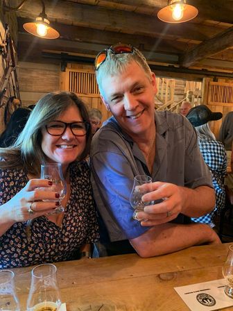 Smiling couple toasting with whiskey tasting glasses at a rustic distillery tasting room, warm wood beams and pendant lights in the background