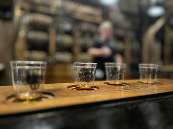 Four-glass whiskey tasting flight on a wooden board with amber samples, set against a blurred distillery barrel room and server in the background.