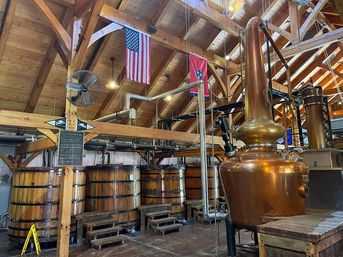Interior of a rustic distillery with a shiny copper pot still, wooden fermentation vats, exposed timber beams, industrial piping, and hanging American and Tennessee flags.