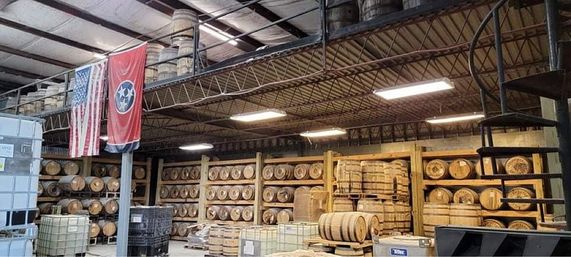 Rustic distillery warehouse interior with rows of stacked wooden whiskey barrels on racks, pallets and tanks on the floor, a spiral metal staircase, and American and Tennessee flags hanging above.