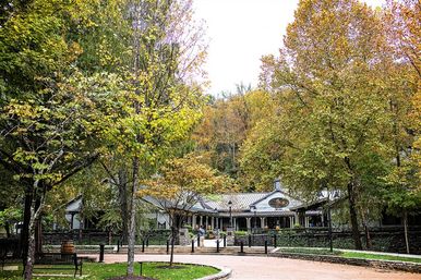 Leafy fall trees frame a quaint pavilion-style building with a covered porch in a mountain park, paved walkway, benches and a wooded hillside.