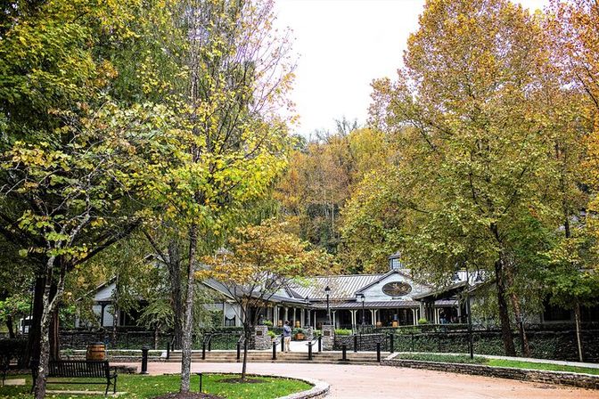 Leafy fall trees frame a quaint pavilion-style building with a covered porch in a mountain park, paved walkway, benches and a wooded hillside.