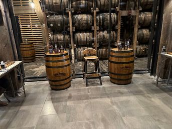 Cozy distillery tasting area with stacked oak whiskey barrels behind glass, two oak barrel tables topped with whiskey bottles and a wooden tasting stool on a tiled floor.