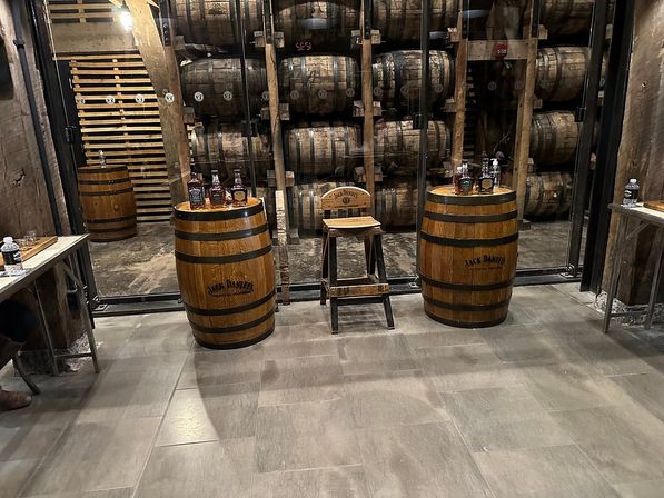 Cozy distillery tasting area with stacked oak whiskey barrels behind glass, two oak barrel tables topped with whiskey bottles and a wooden tasting stool on a tiled floor.