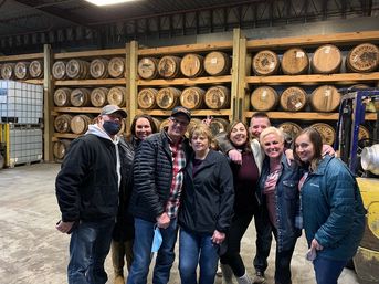 Cheerful group of eight people posing in a distillery warehouse with rows of stacked oak barrels on wooden racks, casual jackets and smiles.