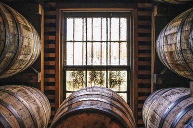 Oak whiskey barrels stacked in a dim distillery aging room around a weathered window with sunlight filtering through.